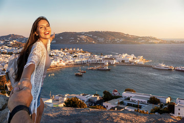 Junge Reisende Frau zieht ihren Partner an der Hand Richtung Mykonos Stadt, Griechenland, während ihres Sommerurlaubes - obrazy, fototapety, plakaty