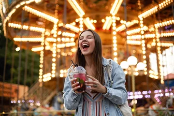 Vlies Fototapete Vergnügungspark Außenporträt einer fröhlichen jungen hübschen brünetten Frau in Freizeitkleidung, die mit geschlossenen Augen und breitem Lächeln über einem Vergnügungspark posiert und eine Tasse Limonade in den Händen hält