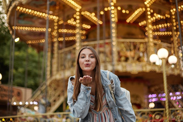 Fototapeta na wymiar Outdoor photo of charming long haired young lady in trendy jeans coat raising palm and blowing air kiss to camera, standing over carousel in park of attractions