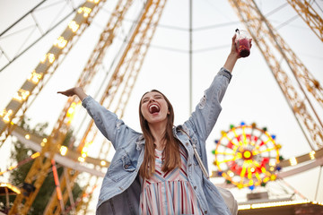 Young joyful long haired pretty female standing over ferris wheel in amusement park, raising hands up and shouting happily with closed eyes, wearing casual clothes