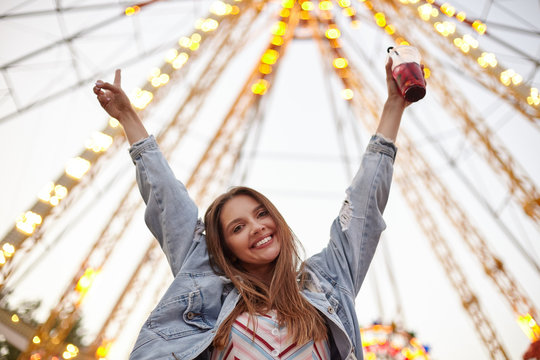 Portrait of beautiful young woman with long brown hair looking to camera happily, posing over attractions in city park with raised hands, wearing casual clothes