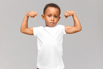 Picture of cute dark skinned little boy in white t-shirt tensing bicep, showing arm muscles, being proud of himself after physical training, looking at camera with confident facial expression