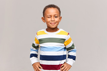 True emotions and body langauge. Studio shot of adorable cute African American little boy dressed in colorful sweater winking at camera with one eye, having mysterious smile, being in good mood