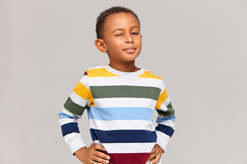 You know what am I saying? Positive confident playful Afro American little boy in multicolored sweater keeping hands on his waist and winking at camera. Human facial expressions and attitude