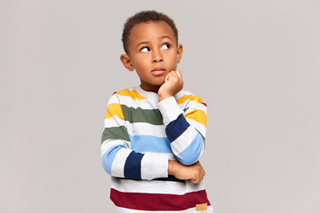 Studio picture of cute dark skinned nine year old boy keeping hand under his chin and looking up with thoughtful pensive facial expression, having curious look, trying to remember something © Anatoliy Karlyuk