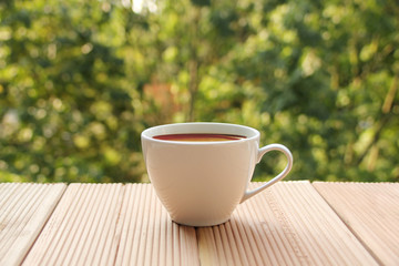 white cup with coffee on a light wooden table against a green summer garden, close-up, copy space