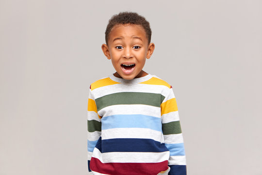 Genuine human facial expressions and feelings. Isolated image of funny cute Afro American boy in striped sweater opening mouth widely and raising eyebrows, being afraid of scary movie or dentist