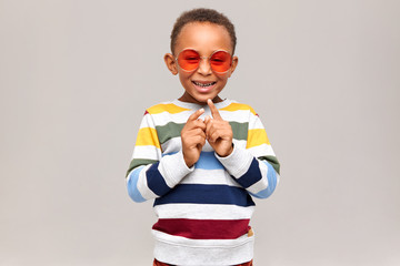 Fashionable adorable African boy in trendy round sunglasses and colorful pullover laughing and...