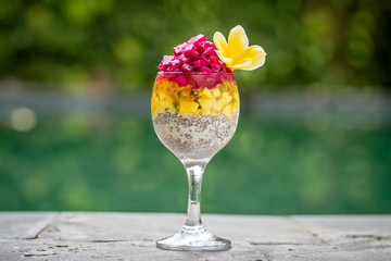 Chia seeds pudding with red dragon fruit, passion fruit, mango and avocado in a glass for breakfast on the background of the swimming pool water, closeup. The concept of healthy eating.