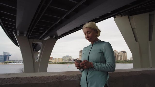 Pan shot of middle-aged blonde Caucasian woman wearing sport clothes standing outdoors under bridge with telephone in her hands and tapping on screenPan shot of middle-aged blonde Caucasian woman wear