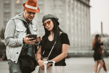 Trendy two hipster people on city street. Stylish couple looking on telephone to find apartment on buildings background. Attractive asian woman and handsome man in hat