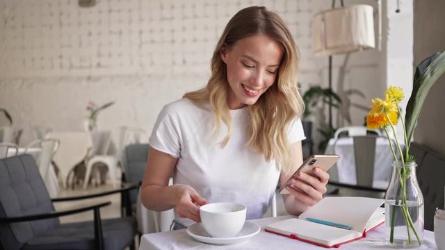Attractive cheerful young blonde woman drinking a cup of coffee and using smartphone while sitting in cafe