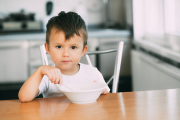 A little boy in the kitchen eating oatmeal from a white plate