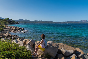 Woman in a dress and with a backpack back view sitting on the rocks on the beach against the mountains