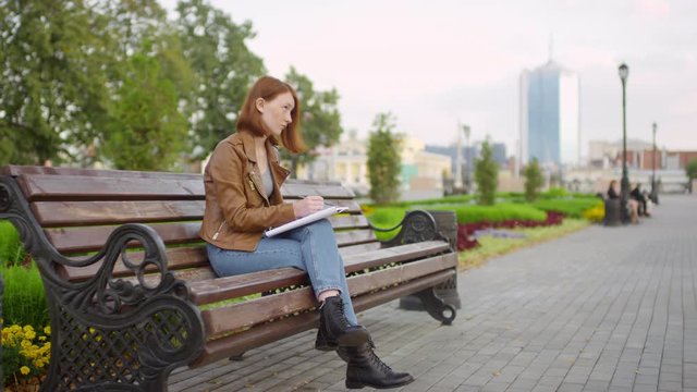 Slowmo tracking shot of beautiful teenage girl with red hair and freckles sitting on bench in city and drawing in sketchbook