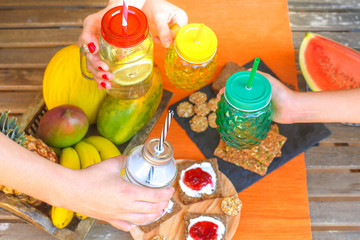 Obraz na płótnie Canvas Hands toasting with natural drinks in colorful glasses with straws. Wooden table with natural food, in background for a healthy and nutritious breakfast. Vegan, vegetarian, bio, diet, meal concept