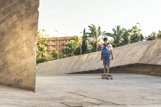 Young Boy Riding A Skate Out Of School In A Urban Context. Teen With Backpack And Rapper Hat, Having Fun With Skateboard In A Modern City.   Happy Student Backing Home From The Institute Youth Concept