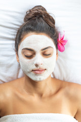 Nice relaxing young woman with white mask on face lying down on towel on bench spa with flower on hair. Smiling on camera.