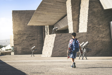 Back view of a young boy with backpack and skateboard in hand in a urban context. Trendy student walking out of the college after a day of school. Youth, new generation, leisure and free time concept