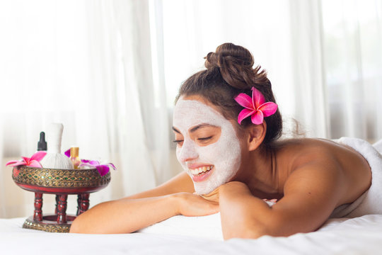 Nice relaxing young woman with white mask on face lying down on towel on bench spa with flower on hair. Smiling on camera.
