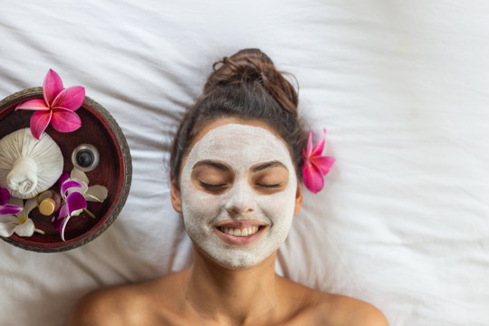 Nice relaxing young woman with white mask on face lying down on towel on bench spa with flower on hair. Smiling on camera.