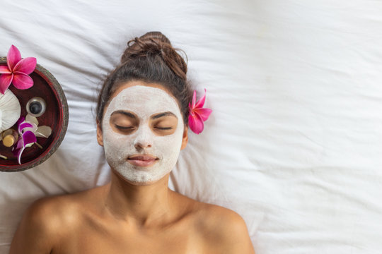 Nice relaxing young woman with white mask on face lying down on towel on bench spa with flower on hair. Smiling on camera.