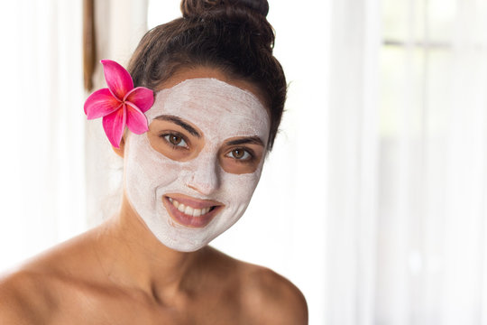 Nice relaxing young woman with white mask on face lying down on towel on bench spa with flower on hair. Smiling on camera.