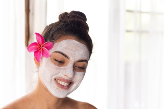 Nice relaxing young woman with white mask on face lying down on towel on bench spa with flower on hair. Smiling on camera.