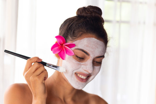 Nice relaxing young woman with white mask on face lying down on towel on bench spa with flower on hair. Smiling on camera.