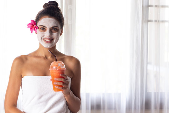 Nice relaxing young woman with white mask on face lying down on towel on bench spa with flower on hair. Smiling on camera.