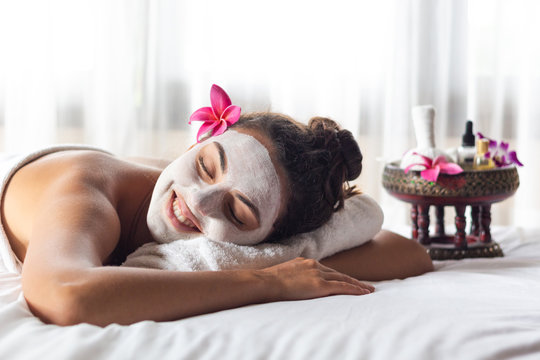 Nice relaxing young woman with white mask on face lying down on towel on bench spa with flower on hair. Smiling on camera.