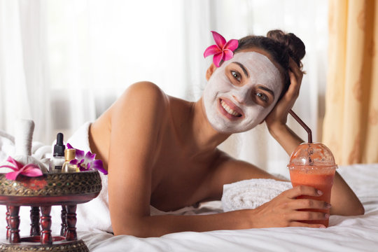 Nice relaxing young woman with white mask on face lying down on towel on bench spa with flower on hair. Smiling on camera.