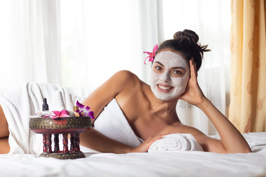 Nice relaxing young woman with white mask on face lying down on towel on bench spa with flower on hair. Smiling on camera.