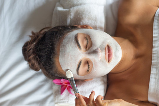 Nice relaxing young woman with white mask on face lying down on towel on bench spa with flower on hair. Smiling on camera.
