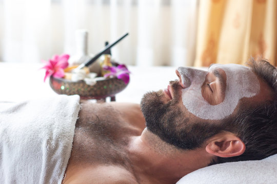 Nice relaxing men with white mask on face lying down on towel on bench spa with flower.