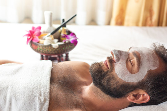 Nice relaxing men with white mask on face lying down on towel on bench spa with flower.