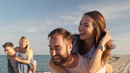 Two cheerful young lovely couples having fun while piggybacking on the beach near the sea - Powered by Adobe