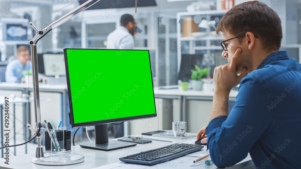 Poster Over the Shoulder Shot of Engineer Working with Green Mock-up Screen Desktop Computer. In the Background Engineering Facility with Specialists Working on Blueprints and Drawings with Industrial Design