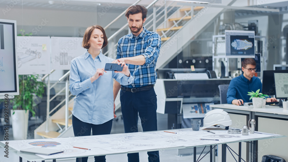 Poster In the Industrial Engineering Facility: Female Designer with Chief Engineer Uses Smartphone for Augmented Reality App to Manipulate Engine Design Technical Drafts in 3D. High Tech Office. Mock-up