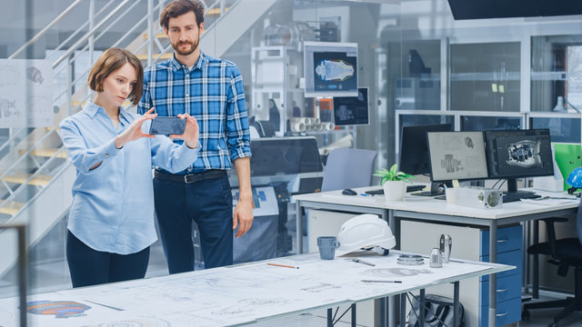 In the Industrial Engineering Facility: Female Designer with Chief Engineer Uses Smartphone for Augmented Reality App to Manipulate Engine Design Technical Drafts in 3D. High Tech Office. Mock-up