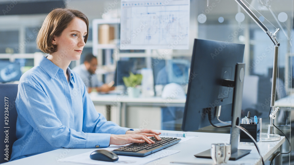 Poster In the Industrial Engineering Facility: Portrait of the Smart and Beautiful Female Engineer Working on Desktop Computer. In the Background Specialists and Technicians Working with Drafts and Drawings