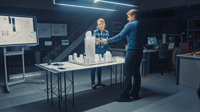 Late at Night in Architectural Agency, Team of Architects and Engineers Working on a City District Buildings Model Prototype. They Talk, Use Digital Tablet. Colleague Works on Desktop Computers