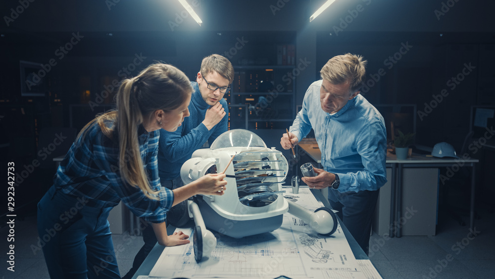 Leinwandbilder Late at Night in Robotics Engineering Facility Three Technical Engineers Talk and Work on a Wheeled Robot Prototype. In the Background High Tech Research Center with Screens Showing Industrial Design
