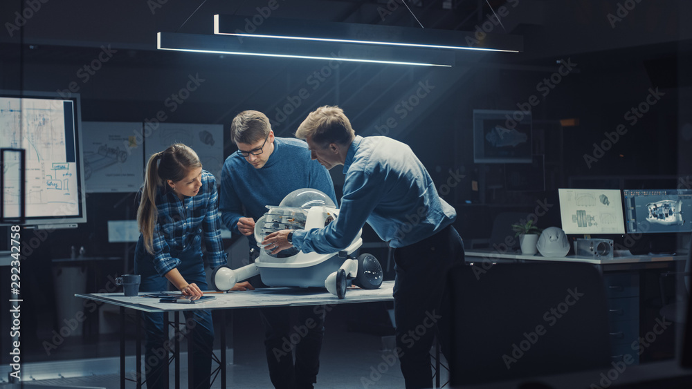 Leinwandbilder Late at Night in Robotics Engineering Facility Three Technical Engineers Talk and Work on a Wheeled Robot Prototype. In the Background High Tech Research Center with Screens Showing Industrial Design