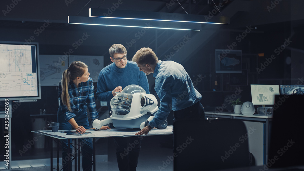 Leinwandbilder Late at Night in Robotics Engineering Facility Three Technical Engineers Talk and Work on a Wheeled Robot Prototype. In the Background High Tech Research Center with Screens Showing Industrial Design