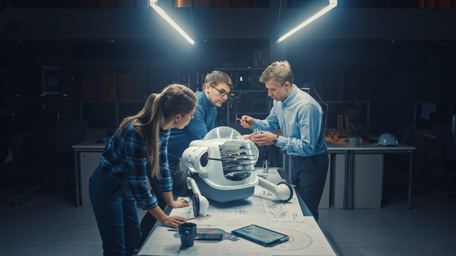 Late at Night in Robotics Engineering Facility Three Technical Engineers Talk and Work on a Wheeled Robot Prototype. In the Background High Tech Research Center with Screens Showing Industrial Design
