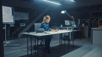 Fototapeta na wymiar Female Engineer Sitting at Her Desk Works with Blueprints Laying on a Table, Uses Pencil, Ruler and Digital Tablet. In the Dark Industrial Design Engineering Facility