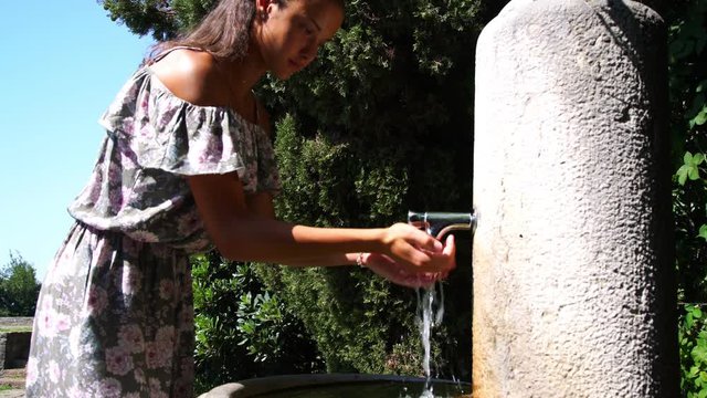 girl in dress with flowers presses tap on old street drinker and drinks water under scorching sun by large tree closeup