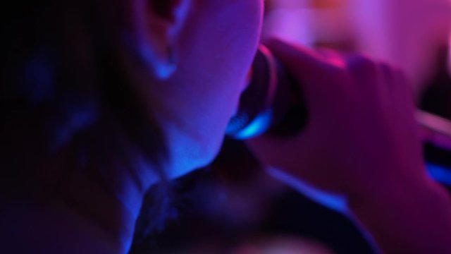 Female vocalist in a hat singing a song in a crowd at a nightclub, close-up. Singer in black perform music at a party. Musician with professional microphone performing on stage under muffled lighting.