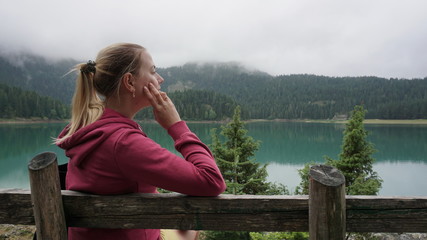 Fototapeta na wymiar Girl sits on a wooden bench in a pink sweatshirt and looks at the black lake, close-up